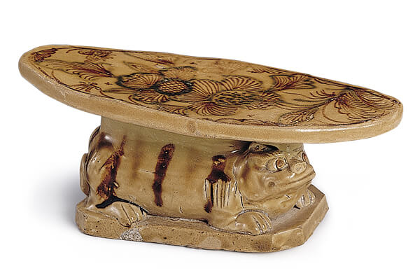 唐代．长沙窑枕+枕面呈椭圆形，下方有一狮子伏卧在方形板上，表面施青釉与褐彩为饰。（图五）